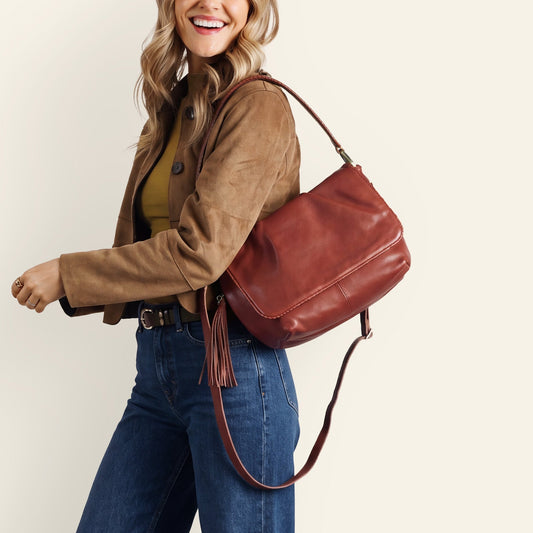 A woman modeling a brown leather shoulder bag with a long strap, styled with a brown jacket, olive top, and wide-leg blue jeans against a solid cream background.