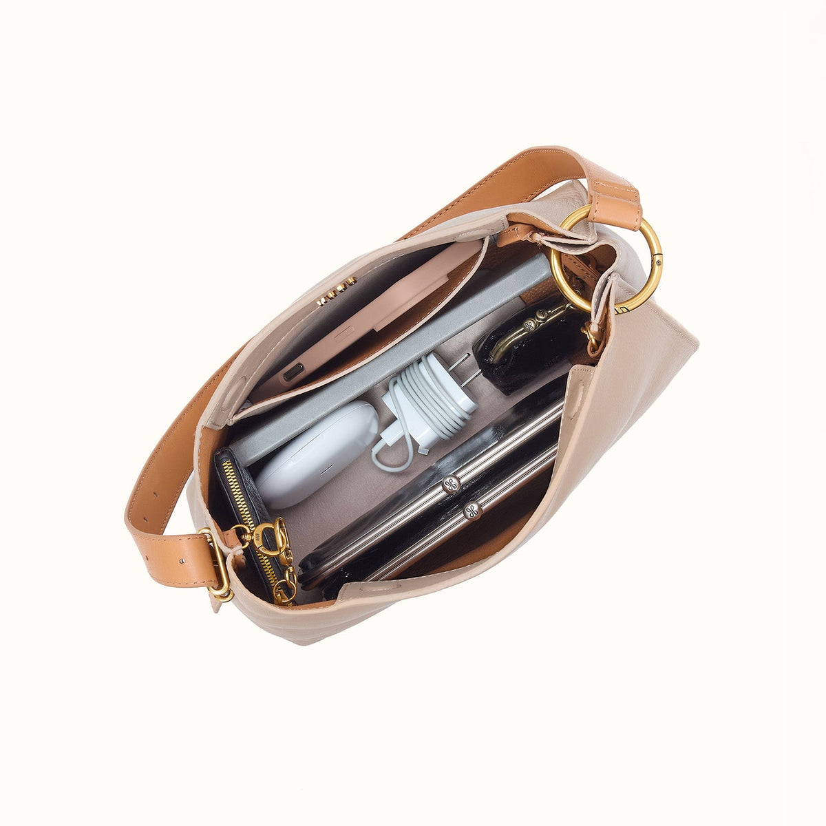 A beige shoulder bag shown open from above, revealing its interior compartments filled with travel accessories including a silver umbrella, white toiletry item, and black wallet. The bag has gold-tone hardware and a tan leather strap on a plain off-white background.